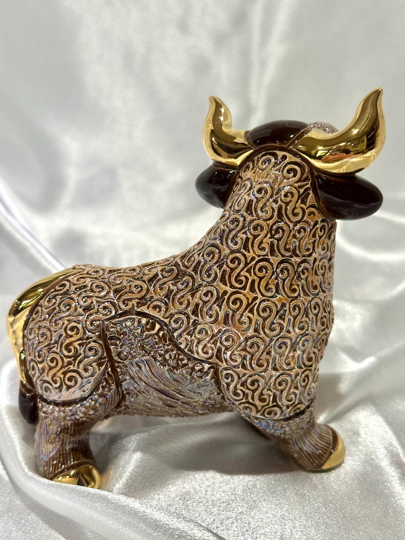 May include: A ceramic bull figurine featuring a detailed brown and tan swirling pattern across its body. The bull has polished gold-colored horns and hooves, with dark brown accents on the ears and head, standing in a profile orientation.