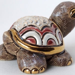 May include: A small ceramic turtle figurine with a brown, white, and red shell. The shell has a gold trim and a detailed design. The turtle is standing on a white surface.