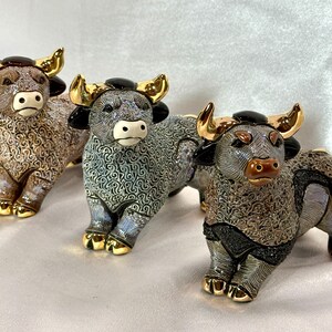 May include: Three ceramic bull figurines featuring intricate swirling patterns on their bodies, glossy black ears, and metallic gold horns and hooves. Each bull is depicted in a standing position with distinct color variations in brown, blue, and grey.