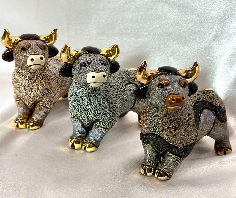 May include: Three ceramic bull figurines featuring intricate swirling patterns on their bodies, glossy black ears, and metallic gold horns and hooves. Each bull is depicted in a standing position with distinct color variations in brown, blue, and grey.