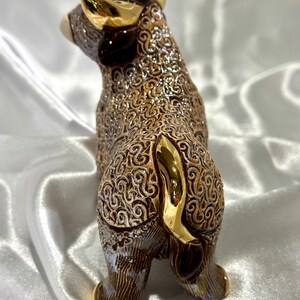May include: A ceramic bull figurine featuring a brown body with an intricate, raised swirling pattern and a metallic, iridescent finish. The bull has polished gold-colored horns and a gold-colored tail, with a smooth, reflective surface texture.