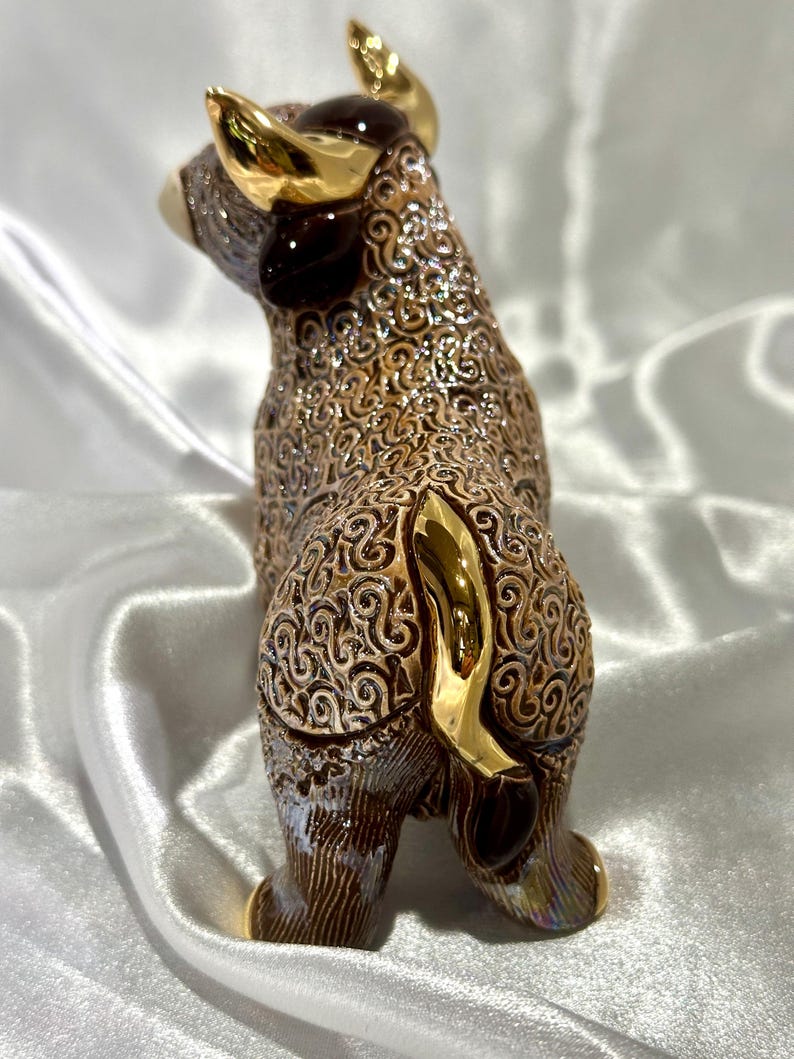 May include: A ceramic bull figurine featuring a brown body with an intricate, raised swirling pattern and a metallic, iridescent finish. The bull has polished gold-colored horns and a gold-colored tail, with a smooth, reflective surface texture.