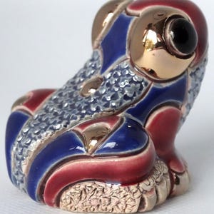 May include: A small ceramic frog figurine with a blue, red, and gold patterned design. The frog has a textured surface and a black eye.