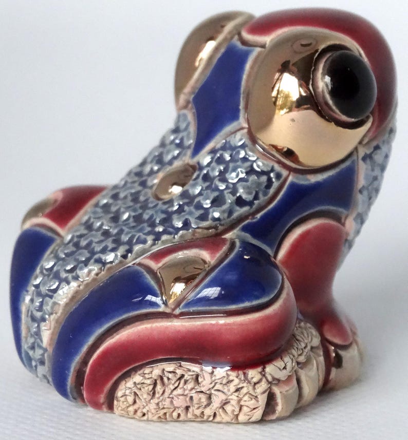 May include: A small ceramic frog figurine with a blue, red, and gold patterned design. The frog has a textured surface and a black eye.