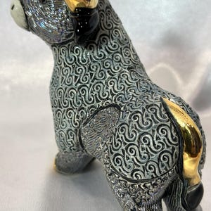 May include: A ceramic bull figurine featuring a textured grey and white swirling pattern across its body. The bull has polished gold-colored horns, a gold-colored tail, and gold-colored hooves, with a smooth white muzzle and black accents.