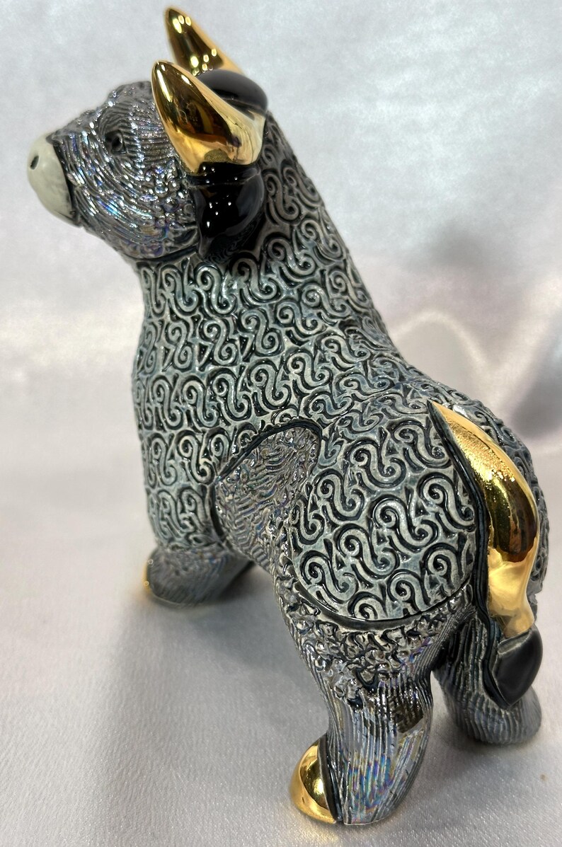 May include: A ceramic bull figurine featuring a textured grey and white swirling pattern across its body. The bull has polished gold-colored horns, a gold-colored tail, and gold-colored hooves, with a smooth white muzzle and black accents.