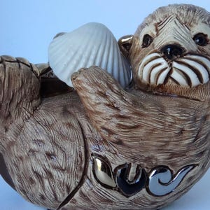 May include: A ceramic figurine of a brown sea otter holding a white seashell. The otter has a detailed face with a white mustache and a gold-colored ear.