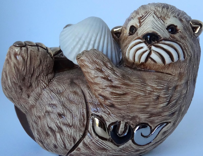 May include: A ceramic figurine of a brown sea otter holding a white seashell. The otter has a detailed face with a white mustache and a gold-colored ear.