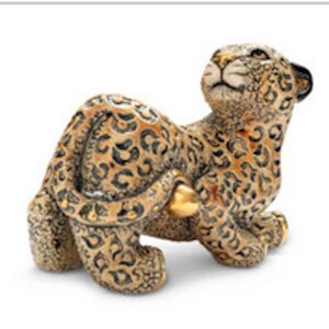 May include: A detailed figurine of a leopard in a resting pose. The leopard has a tan coat with black spots and gold accents. The figurine is looking upwards, with its tail curled around its body.