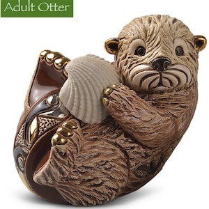 May include: A ceramic figurine of an adult otter holding a seashell. The otter is brown and has a white belly. The seashell is white and has a brown interior. The figurine is sitting on a brown base with gold accents.