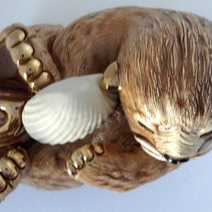 May include: A ceramic figurine of a brown bear holding a white seashell. The bear has gold accents on its paws and ears.