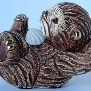 May include: A ceramic figurine of a brown sea otter holding a white seashell. The otter is curled up on its side with its head resting on the shell. The figurine has a realistic appearance with detailed fur and a painted face.