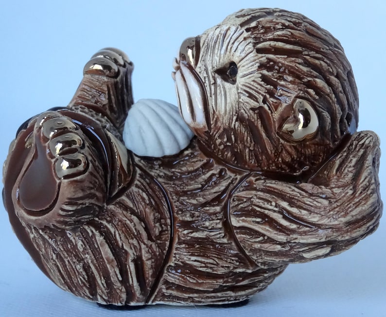 May include: A ceramic figurine of a brown sea otter holding a white seashell. The otter is curled up on its side with its head resting on the shell. The figurine has a realistic appearance with detailed fur and a painted face.