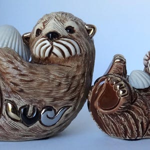 May include: Two ceramic sea otter figurines, one larger than the other, both with brown fur and holding white seashells. The larger otter is lying on its back with its paws in the air, while the smaller otter is sitting up and holding a seashell in its paws. Both otters have realistic details, including their whiskers and eyes.