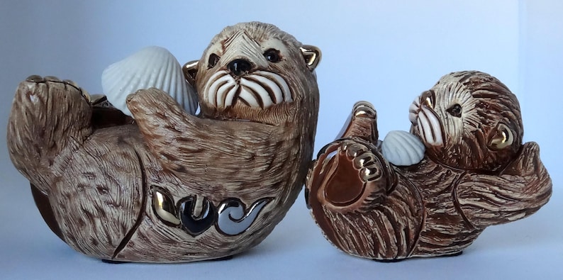 May include: Two ceramic sea otter figurines, one larger than the other, both with brown fur and holding white seashells. The larger otter is lying on its back with its paws in the air, while the smaller otter is sitting up and holding a seashell in its paws. Both otters have realistic details, including their whiskers and eyes.