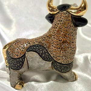 May include: A decorative bull figurine featuring a complex brown and black textured pattern across its body. The bull has polished gold-colored horns and hooves, with smooth black accents on the ears and head, standing in a profile orientation.