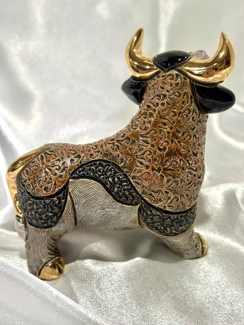 May include: A decorative bull figurine featuring a complex brown and black textured pattern across its body. The bull has polished gold-colored horns and hooves, with smooth black accents on the ears and head, standing in a profile orientation.