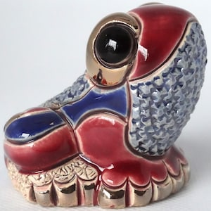 May include: A ceramic frog figurine with a red, blue, and gold glaze. The frog has a large, black eye with a gold rim. The frog is sitting on a base with a textured surface.