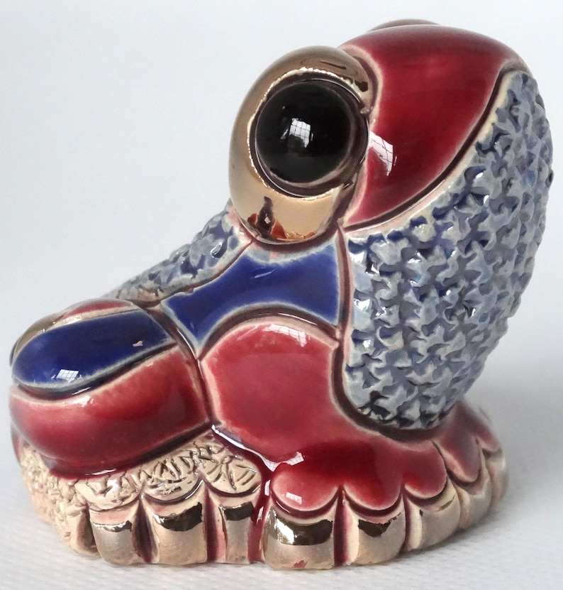 May include: A ceramic frog figurine with a red, blue, and gold glaze. The frog has a large, black eye with a gold rim. The frog is sitting on a base with a textured surface.