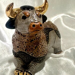 May include: A ceramic bull figurine featuring a textured brown and grey body with intricate swirling patterns. The bull has polished gold-tone horns and hooves, a smooth black patch on its forehead, and a brown snout with two distinct nostrils.