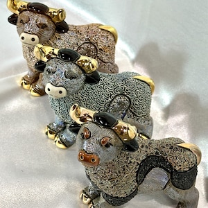 May include: Three ceramic bull figurines featuring intricate, textured patterns across their bodies. Each bull has polished gold-tone horns and hooves. The figures are arranged in a diagonal line, displaying varying color palettes of brown, grey, and black.