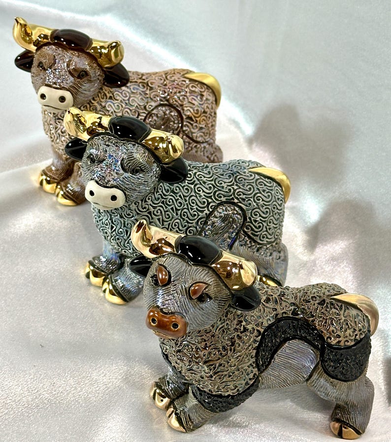 May include: Three ceramic bull figurines featuring intricate, textured patterns across their bodies. Each bull has polished gold-tone horns and hooves. The figures are arranged in a diagonal line, displaying varying color palettes of brown, grey, and black.