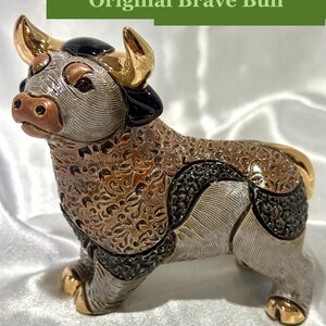May include: A ceramic bull figurine featuring a textured brown and silver body with intricate swirling patterns. The bull has polished gold-colored horns and hooves, with black accents on the head and flanks. The text Original Brave Bull is visible.
