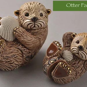 May include: Two ceramic otter figurines, one brown and one tan, holding seashells. The otters are in a playful pose, with their heads tilted and their eyes closed. The figurines are part of an otter family collection.