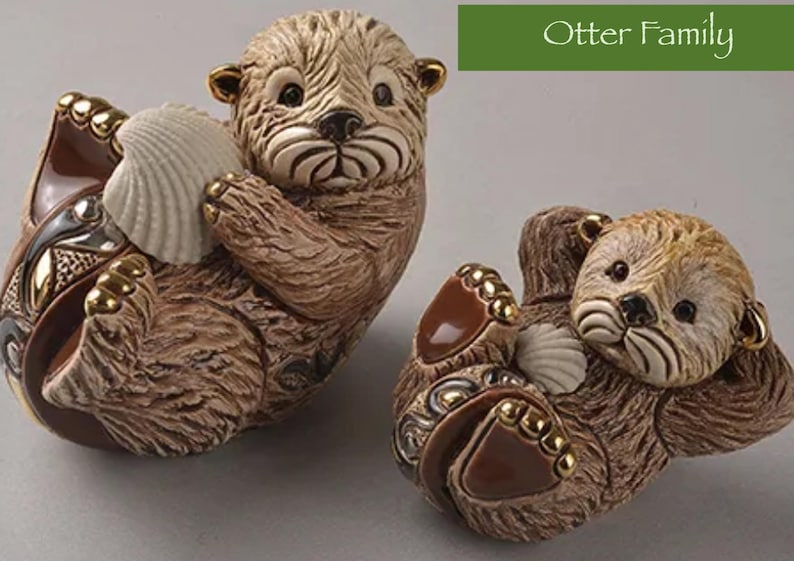 May include: Two ceramic otter figurines, one brown and one tan, holding seashells. The otters are in a playful pose, with their heads tilted and their eyes closed. The figurines are part of an otter family collection.