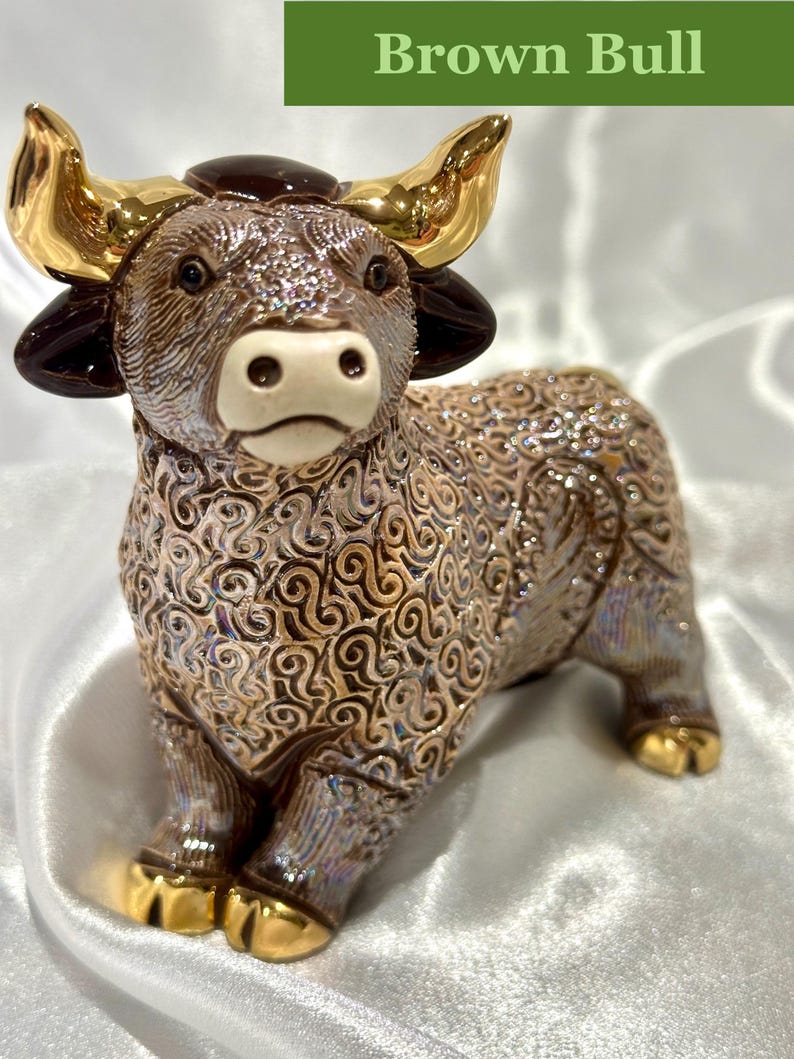 May include: A ceramic bull figurine featuring a textured, iridescent brown body with swirling patterns. The bull has polished gold-colored horns and hooves, dark brown ears, and a cream-colored muzzle. The text "Brown Bull" appears at the top.