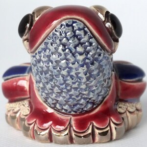 May include: A ceramic frog figurine with a red, blue, and gold glaze. The frog is sitting on its hind legs with its front legs folded in front of it. The frog's eyes are black and the glaze has a textured finish.