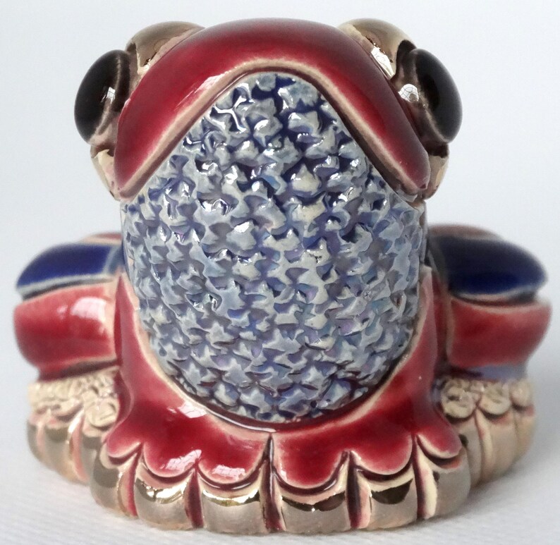 May include: A ceramic frog figurine with a red, blue, and gold glaze. The frog is sitting on its hind legs with its front legs folded in front of it. The frog's eyes are black and the glaze has a textured finish.