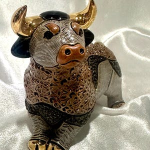 May include: A ceramic bull figurine featuring a textured brown and grey body with intricate swirling patterns. The bull has polished gold-tone horns and hooves, a smooth black patch on its forehead, and a brown snout with two distinct nostrils.