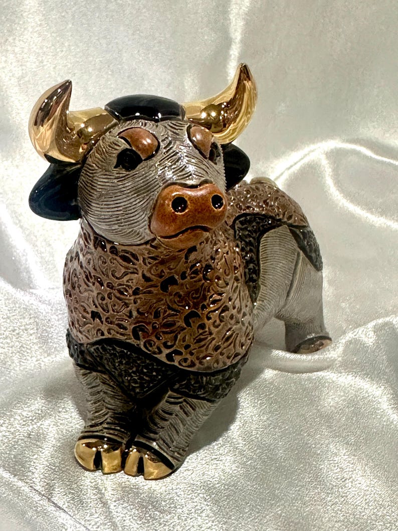 May include: A ceramic bull figurine featuring a textured brown and grey body with intricate swirling patterns. The bull has polished gold-tone horns and hooves, a smooth black patch on its forehead, and a brown snout with two distinct nostrils.