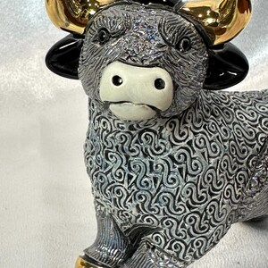 May include: A ceramic bull figurine featuring a textured grey and black body with a swirling pattern. The bull has a smooth white muzzle, glossy black ears, and metallic gold horns and hooves. The surface of the body has a detailed, raised relief design.