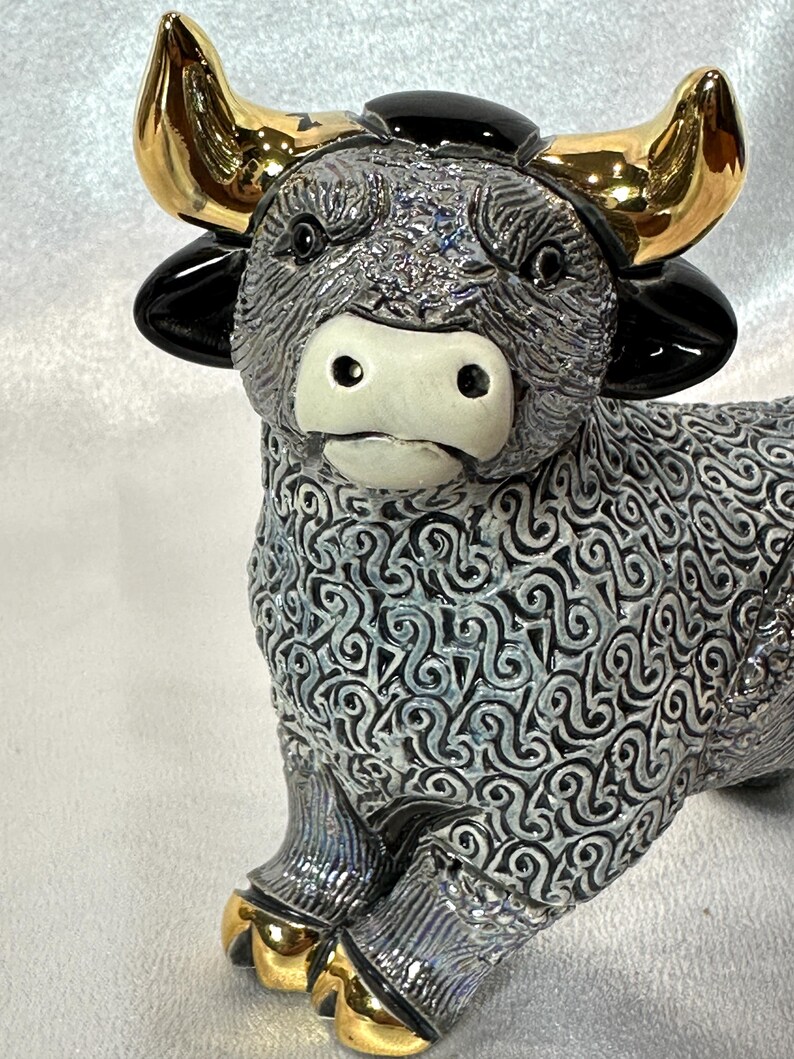 May include: A ceramic bull figurine featuring a textured grey and black body with a swirling pattern. The bull has a smooth white muzzle, glossy black ears, and metallic gold horns and hooves. The surface of the body has a detailed, raised relief design.