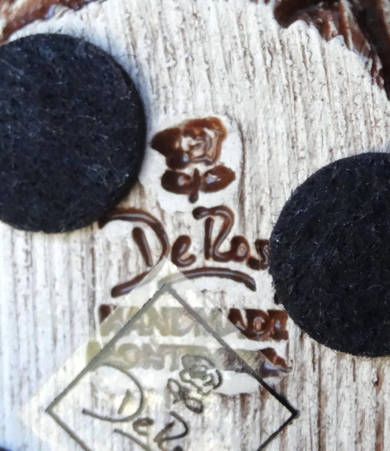 May include: Close-up of a white ceramic figurine with a brown glaze. The figurine has a De Rosa Hand Made Holland label and a rose logo.