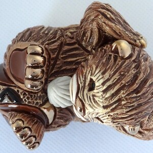 May include: A brown ceramic figurine of a bear cub with gold accents. The bear is lying on its side with its head resting on its paw. The bear has a white, textured object in its mouth.