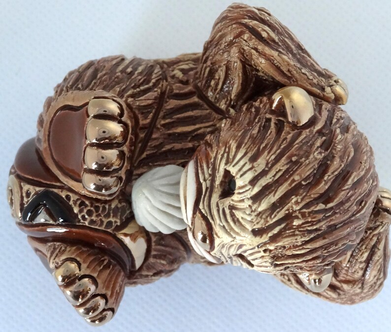 May include: A brown ceramic figurine of a bear cub with gold accents. The bear is lying on its side with its head resting on its paw. The bear has a white, textured object in its mouth.
