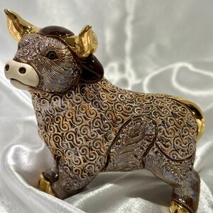 May include: A ceramic bull figurine featuring a textured, swirling brown and iridescent finish. The bull has polished gold-colored horns, hooves, and tail accents, with a smooth, cream-colored muzzle and dark brown ears, standing in a side profile view.