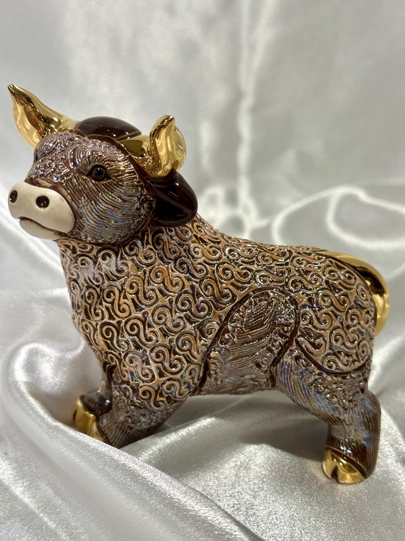 May include: A ceramic bull figurine featuring a textured, swirling brown and iridescent finish. The bull has polished gold-colored horns, hooves, and tail accents, with a smooth, cream-colored muzzle and dark brown ears, standing in a side profile view.