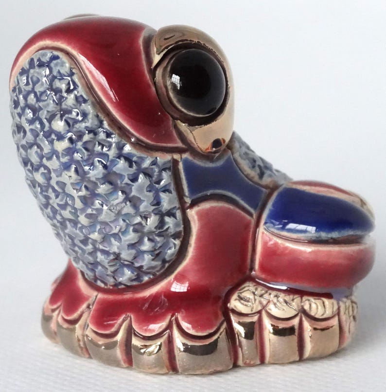 May include: A small ceramic frog figurine with a red, blue, and gold glaze. The frog has a large, black eye and a detailed, textured surface.