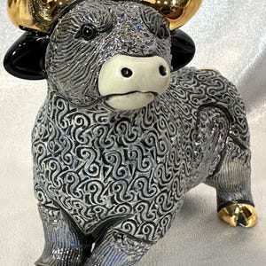 May include: A ceramic bull figurine featuring a textured grey and black swirling pattern across its body. The bull has a smooth white muzzle, glossy black ears, and polished gold-tone horns and hooves. The surface has a reflective, glazed finish.