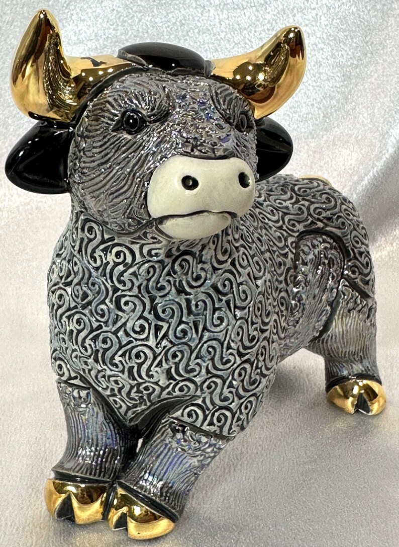 May include: A ceramic bull figurine featuring a textured grey and black swirling pattern across its body. The bull has a smooth white muzzle, glossy black ears, and polished gold-tone horns and hooves. The surface has a reflective, glazed finish.