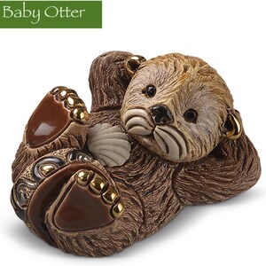 May include: A ceramic figurine of a baby otter with brown fur, lying on its back with its paws outstretched. The otter has a shell in its hand and is holding a small, golden ring in its ear.
