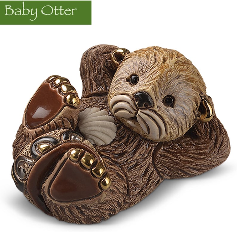 May include: A ceramic figurine of a baby otter with brown fur, lying on its back with its paws outstretched. The otter has a shell in its hand and is holding a small, golden ring in its ear.