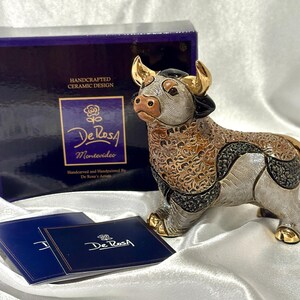 May include: A hand-carved and hand-painted ceramic bull figurine featuring gold-tone horns and hooves, with intricate textured patterns in shades of brown, black, and silver. The bull is displayed with its original dark blue box and informational card.