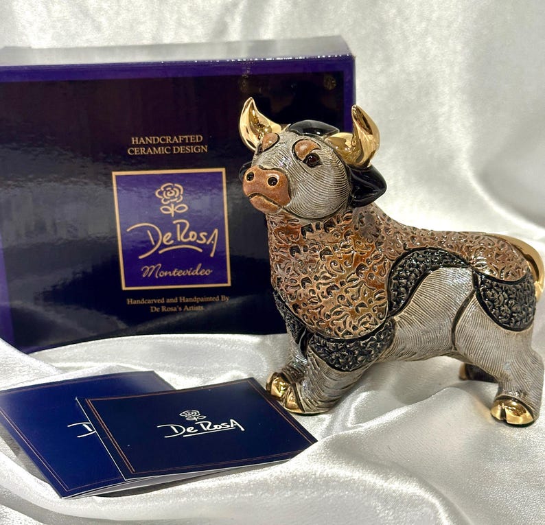 May include: A hand-carved and hand-painted ceramic bull figurine featuring gold-tone horns and hooves, with intricate textured patterns in shades of brown, black, and silver. The bull is displayed with its original dark blue box and informational card.