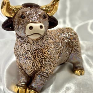 May include: A ceramic bull figurine featuring a textured, iridescent brown body with intricate swirling patterns. The bull has polished gold-colored horns and hooves, dark brown ears, and a smooth, cream-colored muzzle. The surface has a reflective finish.