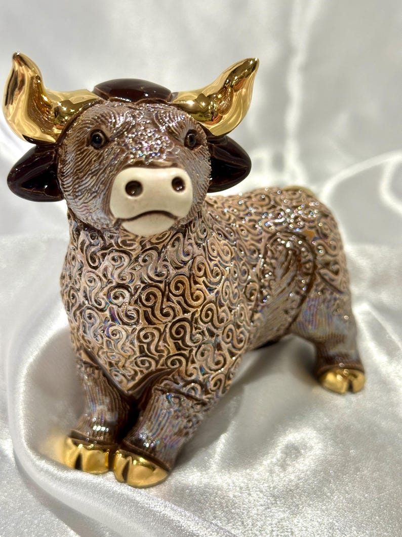 May include: A ceramic bull figurine featuring a textured, iridescent brown body with intricate swirling patterns. The bull has polished gold-colored horns and hooves, dark brown ears, and a smooth, cream-colored muzzle. The surface has a reflective finish.
